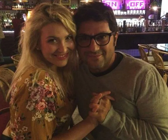 A couple smiling at the camera, holding hands, seated in a dimly lit bar or restaurant.
