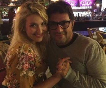 A couple smiling at the camera, holding hands, seated in a dimly lit bar or restaurant.