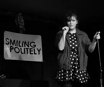 Comedian on stage with a microphone, wearing a polka dot dress. A sign reads "Smiling Politely".