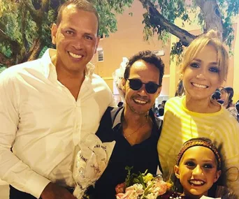 Family smiling outdoors: two adults, one child with flowers, and another person in sunglasses, tree in background.
