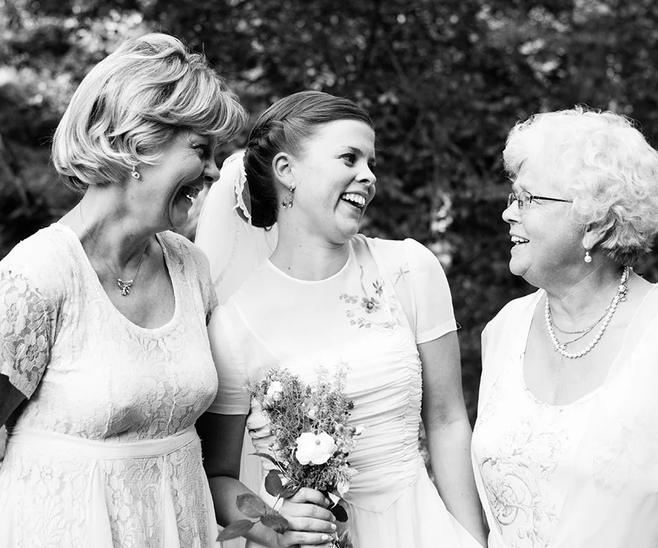 Bride laughing with two older women in a garden, all wearing dresses.