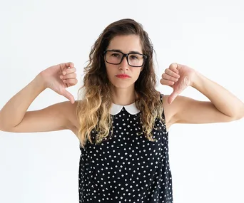 Woman with glasses giving thumbs down, wearing a polka dot dress against a plain background.