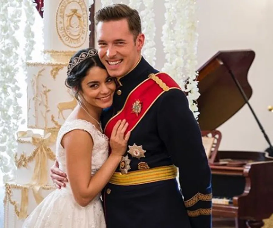 A couple in wedding attire smiles warmly, standing close together in front of a floral-decorated pillar indoors.