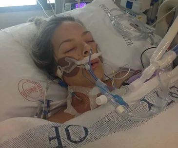 A woman in a hospital bed with tubes and medical equipment, appearing unconscious.
