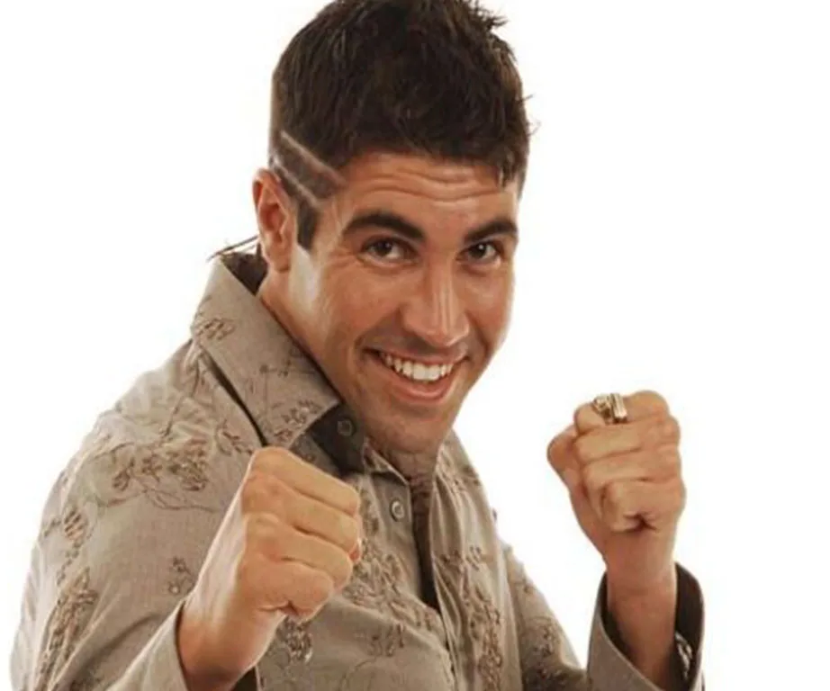 A man with spiked hair, smiling, wearing a patterned shirt, and playfully posing with clenched fists.