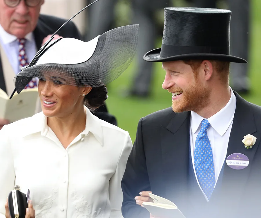 A couple in formal attire smiles at Royal Ascot 2018. The woman wears a large hat, and the man wears a top hat.