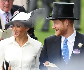 A couple in formal attire smiles at Royal Ascot 2018. The woman wears a large hat, and the man wears a top hat.