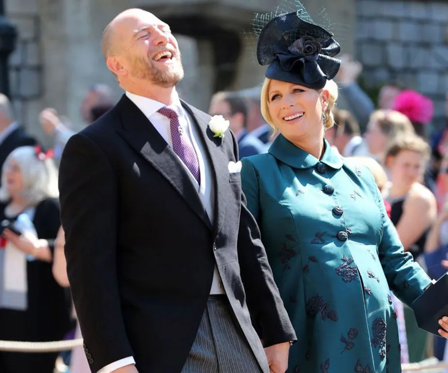 A smiling couple dressed formally at an outdoor event; man in a suit and woman in a dark teal coat with a hat.