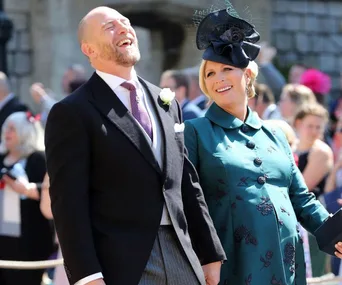 A smiling couple dressed formally at an outdoor event; man in a suit and woman in a dark teal coat with a hat.