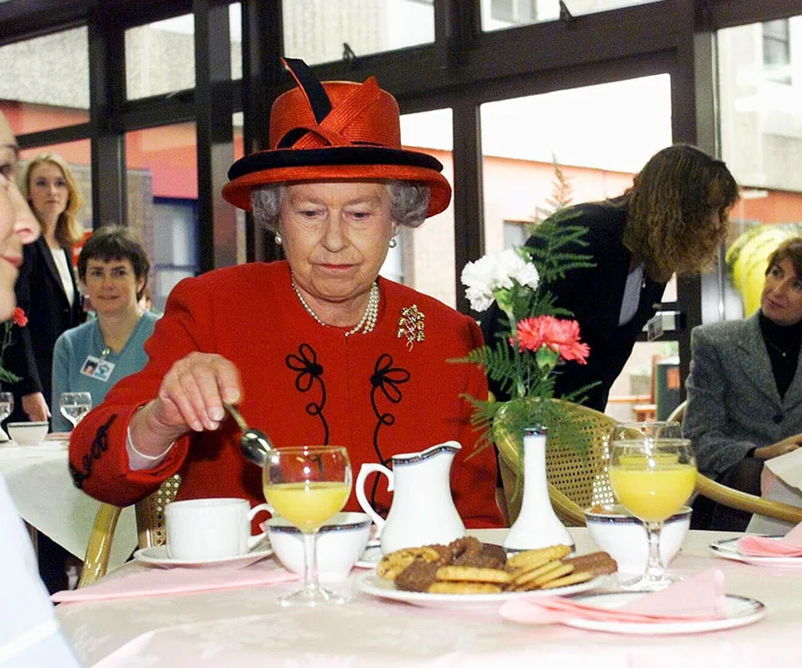 A woman in a red outfit and hat pours juice at a table set with cookies and flowers, surrounded by several people.
