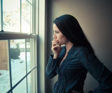 A woman stands by a window, looking outside thoughtfully, with her hand on her chin. Snow is visible outside.