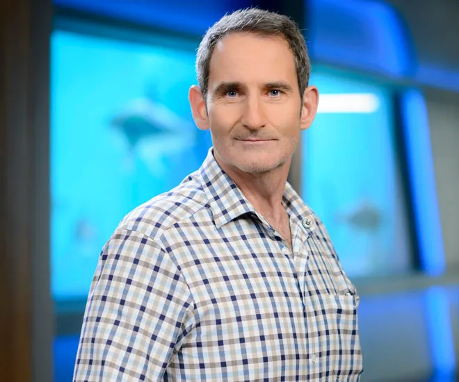 A person in a checkered shirt standing in front of a blurred aquatic-themed background.