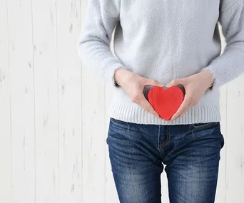 Person in sweater and jeans holding a red heart near their abdomen, symbolizing gut health.