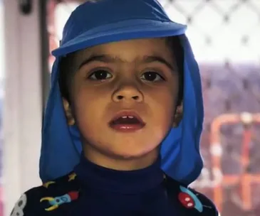 A young boy wearing a blue sunhat and colorful top stands indoors, gazing ahead with a serious expression.