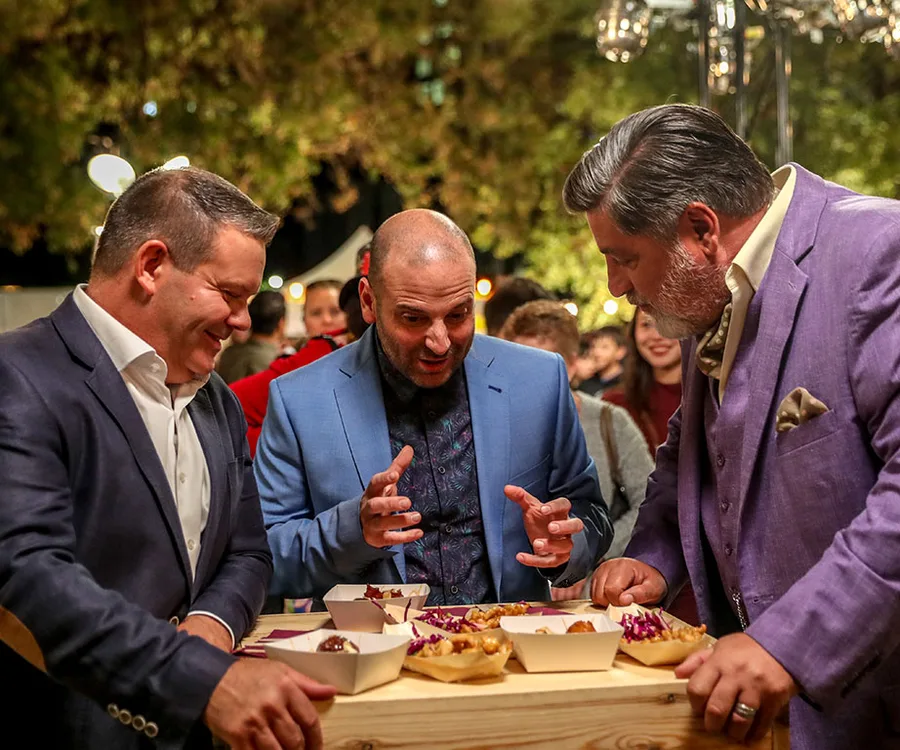 Three judges at a food event, discussing dishes served in cardboard trays, with an outdoor setting in the background.