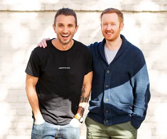 Two smiling men stand together; one in a black T-shirt and jeans, the other in a navy cardigan; outdoor background.