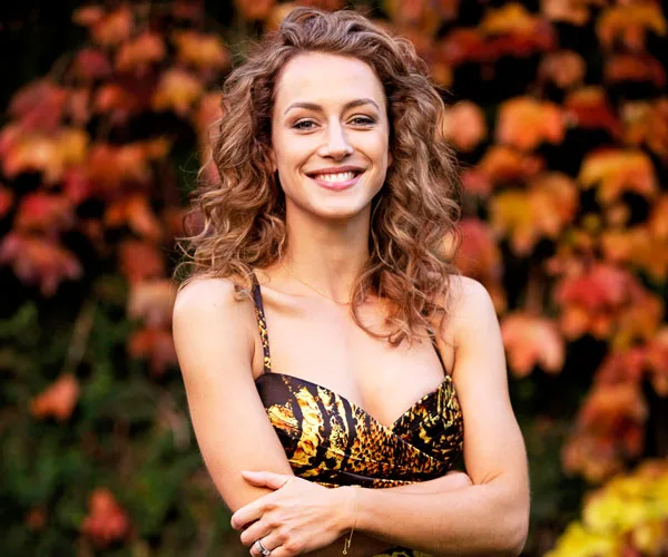 Smiling woman with curly hair wearing a patterned dress, standing in front of autumnal foliage backdrop.