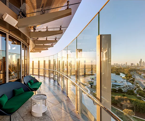 Luxury hotel balcony with glass railing overlooking city skyline and river at sunset, featuring modern chairs and decor.