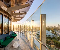 Luxury hotel balcony with glass railing overlooking city skyline and river at sunset, featuring modern chairs and decor.