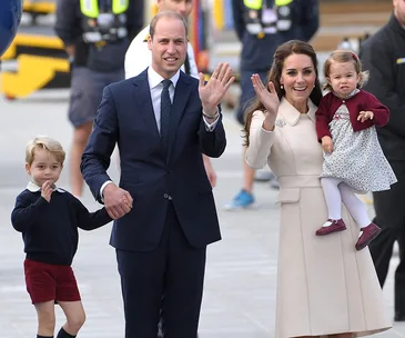 A family waves at an event, the parents in formal attire, with their two young children, one held in the mother's arms.