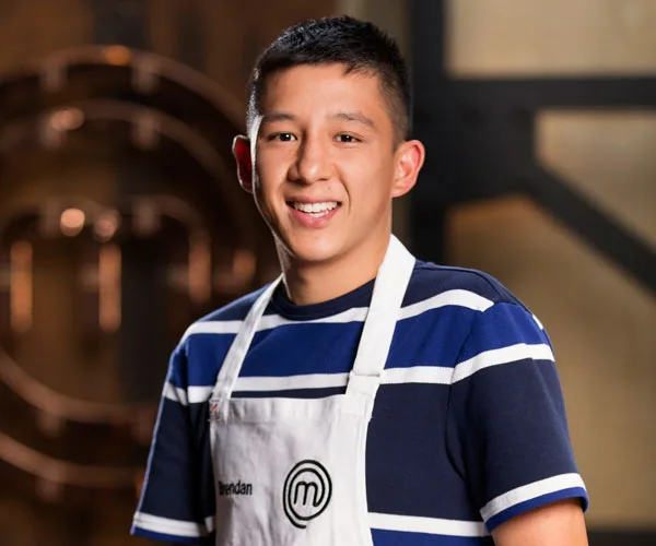 Chef in white apron and striped shirt smiles in a kitchen setting on "MasterChef" show.
