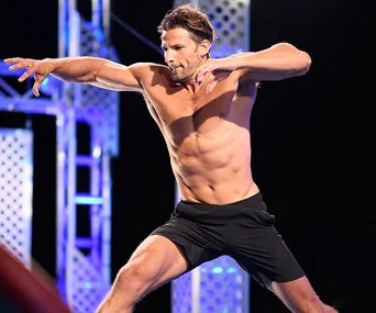 Man in athletic shorts performing an action pose on "Ninja Warrior" set with metal structures in the background.