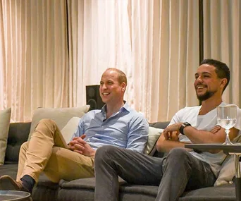 Two men sitting on a couch, smiling and relaxed, with a glass on the table in front of them.