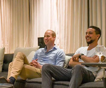 Two men sitting on a couch, smiling and relaxed, with a glass on the table in front of them.