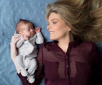 Mother smiling at her sleeping baby, both lying on a blue blanket.