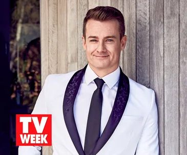 Man in a white suit with black tie leaning against a wall, smiling. TV WEEK logo in bottom corner.