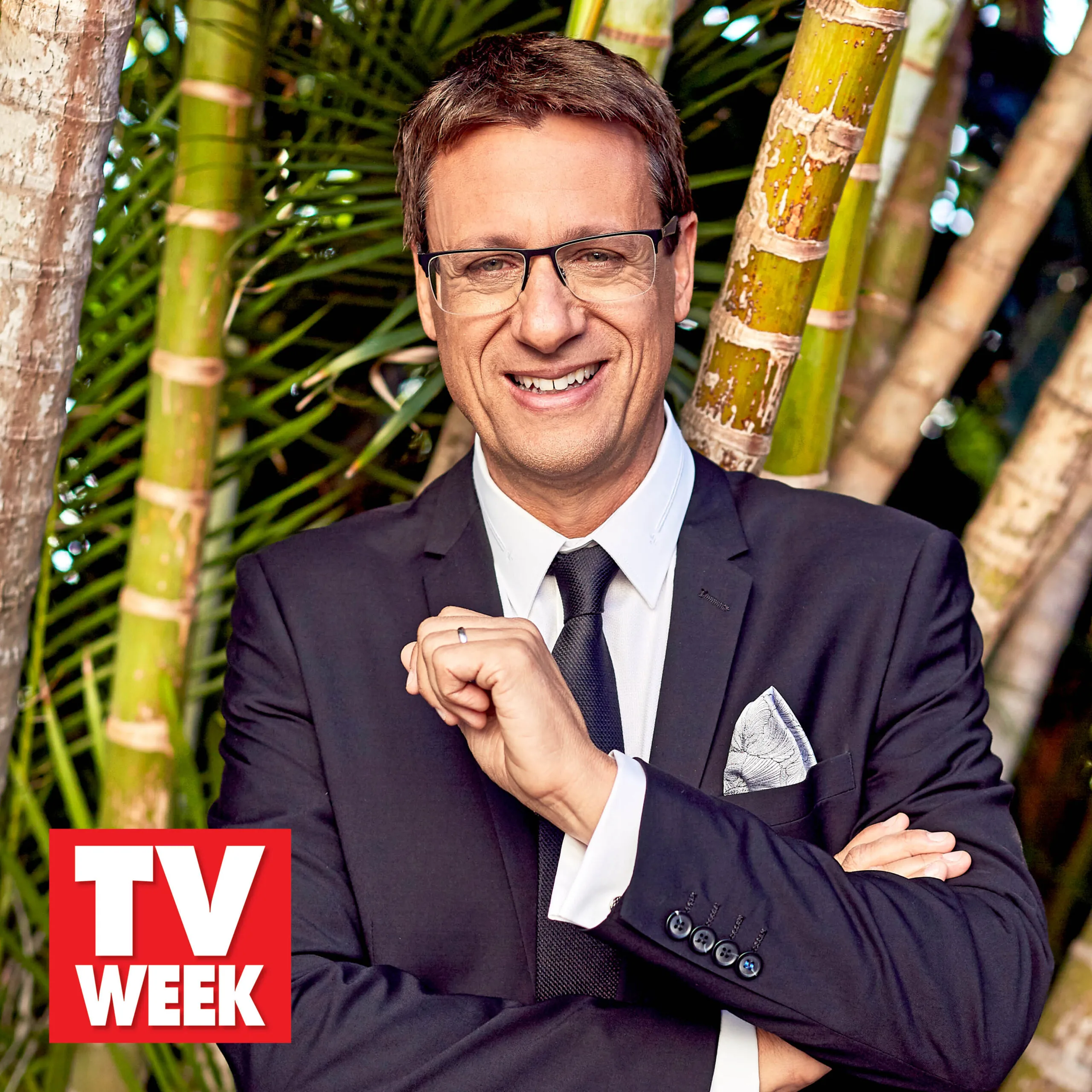 Man in suit smiling, wearing glasses, with crossed arms, against a backdrop of bamboo, "TV Week" logo in corner.