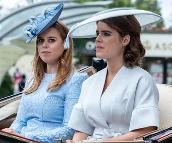 Two women in formal blue outfits and hats sit side by side in a carriage at an outdoor event.