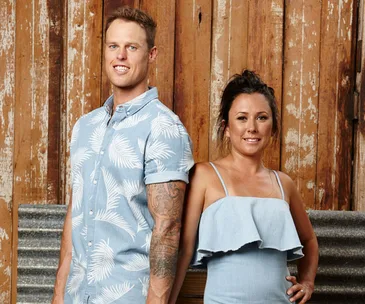 Couple standing with rustic wooden background, both in light blue outfits, smiling.
