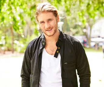 Man in a black jacket over a white shirt standing outdoors, smiling.