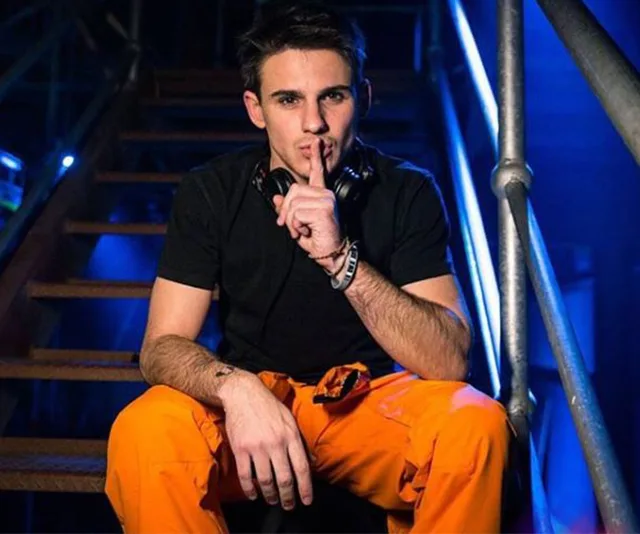 Young man in black shirt and orange pants sitting on stairs, holding finger to lips in a shushing gesture.