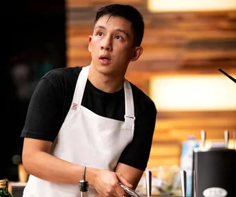 A contestant in a white apron appears focused during a cooking competition.
