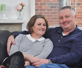 A couple sitting closely on a sofa, smiling; a small flower vase visible in the background.