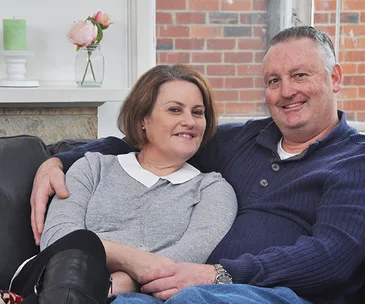 A couple sitting closely on a sofa, smiling; a small flower vase visible in the background.