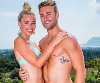 Couple smiling and embracing in swimwear with scenic mountain and ocean backdrop.