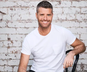 Person smiling, seated against a weathered brick wall, wearing a white t-shirt.