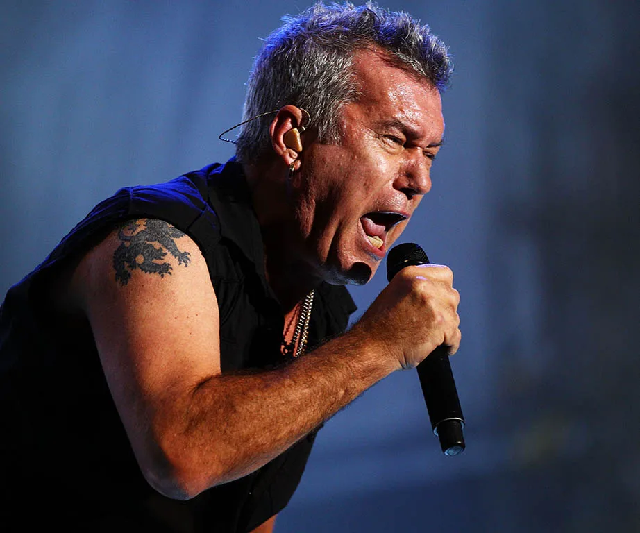 Rock singer passionately performing on stage, gripping microphone, with visible tattoo on upper arm.