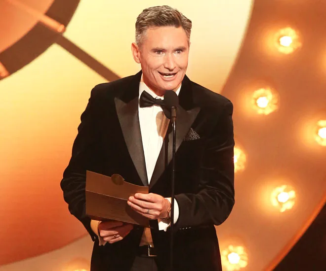 Dave Hughes in a black tuxedo speaks at the 2018 Logies, holding an envelope on stage with a golden backdrop.