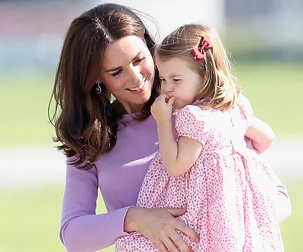 Mother in a lavender dress holds her young daughter wearing a pink dress with a bow, smiling warmly outdoors.