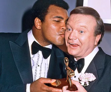 Bert Newton and Muhammad Ali at the 1979 Logies, with Ali playfully kissing Newton on the cheek, both in tuxedos.