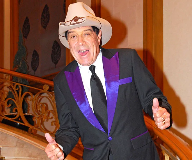 Man in a suit with purple trim, wearing a cowboy hat, giving thumbs up on a decorative staircase.