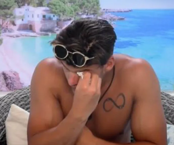 A shirtless man with sunglasses on his head wipes his eye, sitting outdoors with a beach backdrop.