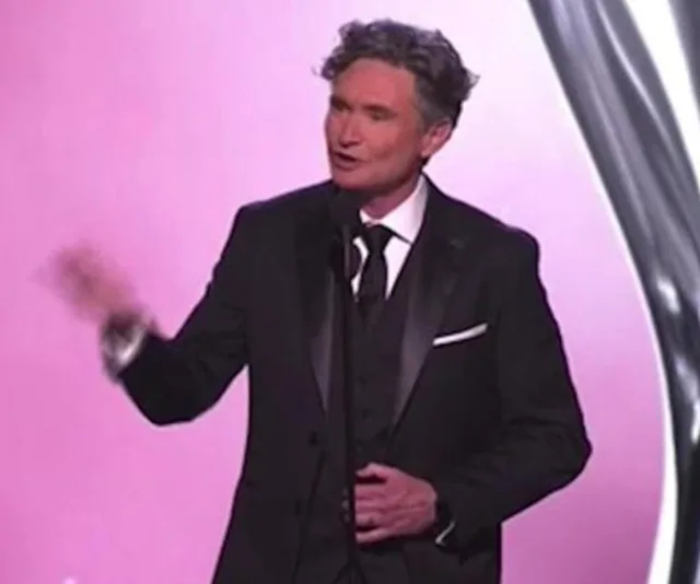 Man in a tuxedo speaking on stage with a microphone during an event with a pink backdrop.