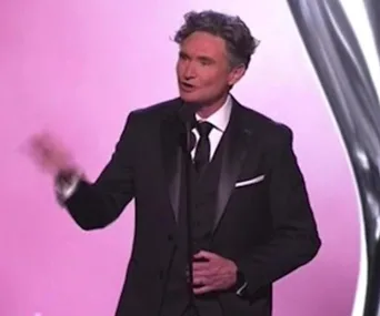Man in a tuxedo speaking on stage with a microphone during an event with a pink backdrop.