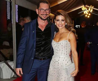 Man in blue suit with glasses and woman in a white dress posing together on a red carpet.