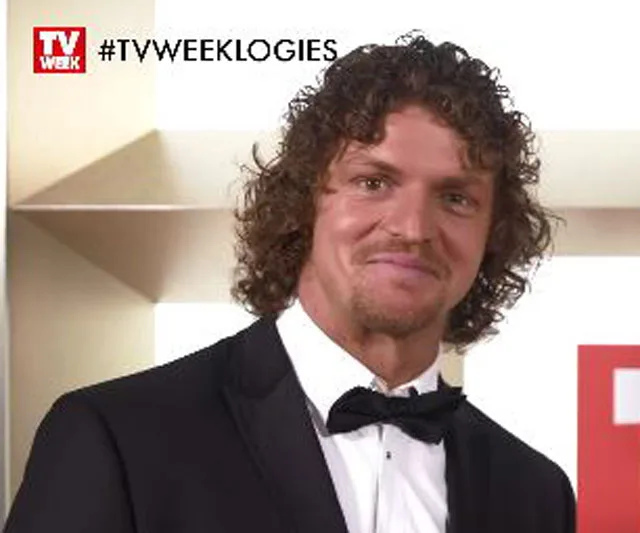 Man in a tuxedo smiling at a TV event backdrop with "TV WEEK LOGIES" hashtag in top corner.
